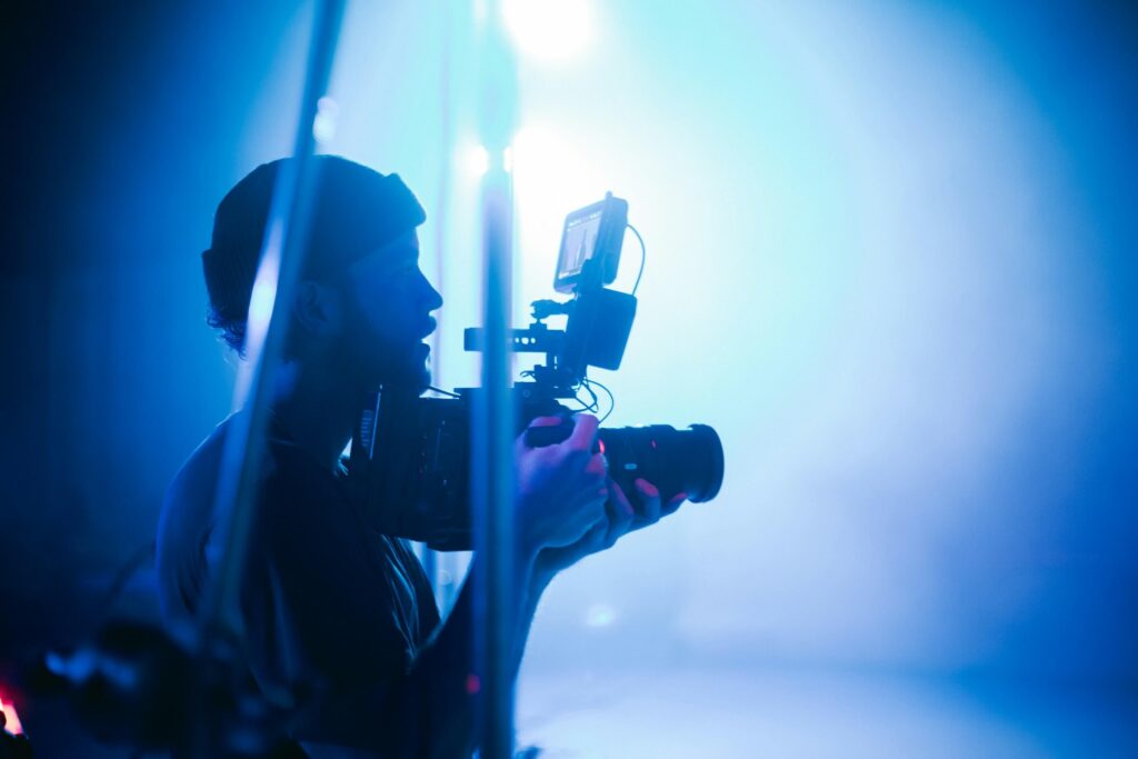 Camera operator filming a live event with a professional video camera, silhouetted against bright blue stage lighting