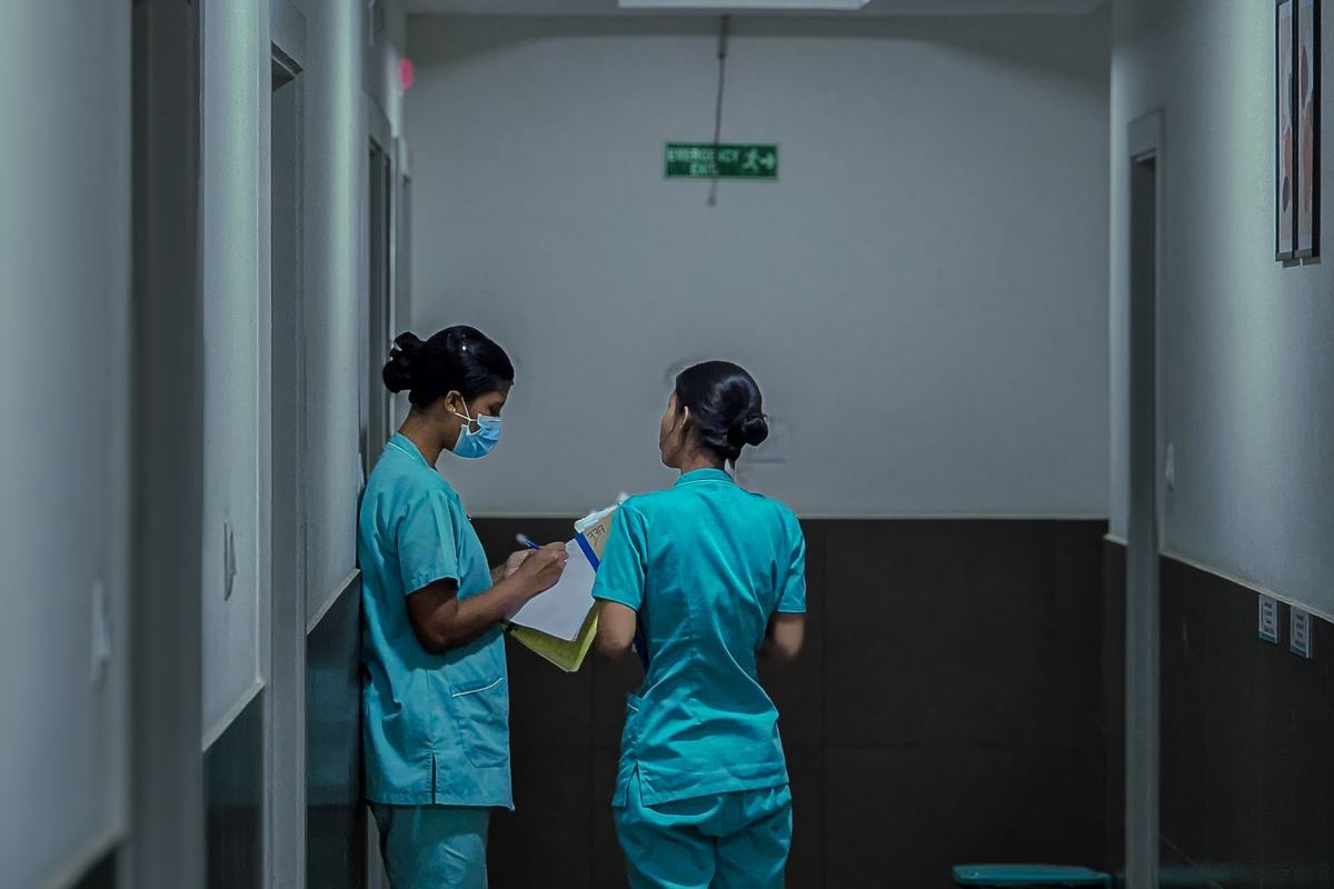 Two nurses talking in a hospital corridor, one writing on a clipboard